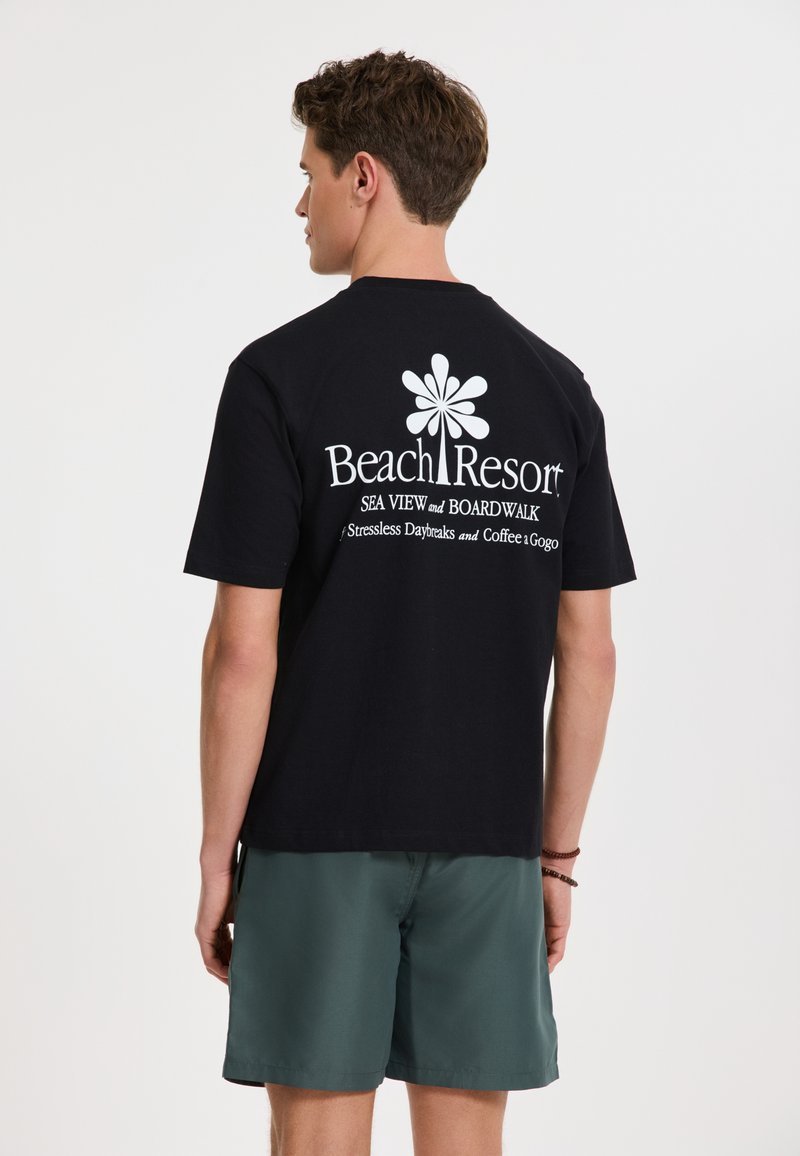Juodas medvilninis marškinėliai su balta užrašu "Beach Resort", su gėlių grafika ir dizainu nugaroje. Dėvimi su žaliais šortais.