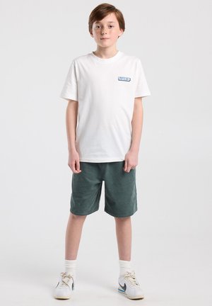 Jongen met lichtbruin haar staat naar voren kijkend en draagt een wit T-shirt met "SURF" opdruk, donkergroene korte broek, witte sokken en witte Nike sneakers.