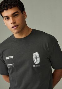 Tricou din bumbac gri închis, având text grafic alb și un design circular pe față. Gât rotund și mâneci scurte cu textură netedă.