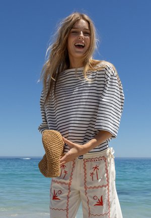 Jeune femme souriante au bord de la mer portant une chemise rayée et un pantalon blanc avec des motifs de palmiers rouges, tenant un chapeau de paille tressé.