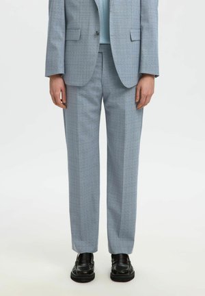 Person iført lysegrå skræddersyede bukser, matchende blazer, lyseblå top og sorte læder loafers, stående mod hvid baggrund.