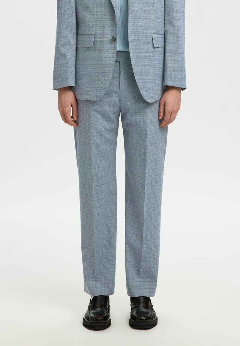 Personne portant un pantalon de costume gris clair sur mesure, un blazer assorti, un haut bleu clair et des mocassins en cuir noirs, debout devant un fond blanc.