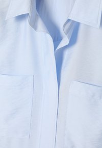 Svijetloplava košulja s klasičnom ovratnikom, dva prednja džepa i glatkom teksturom tkanine s suptilnim detaljima šivanja.