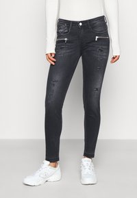 Jeans ajustados negros con detalles desgastados, bolsillos con cremallera y diseño estándar de cinco bolsillos. Llevados con zapatillas deportivas blancas.