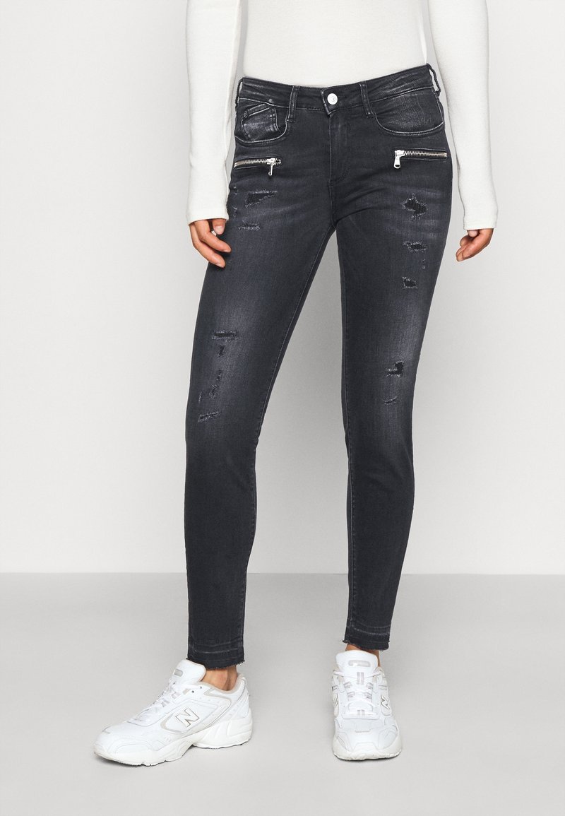 Jeans ajustados negros con detalles desgastados, bolsillos con cremallera y diseño estándar de cinco bolsillos. Llevados con zapatillas deportivas blancas.