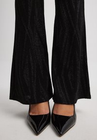 Zwarte flared broeken hebben een gestructureerd patroon met glanzende accenten; gecombineerd met elegante zwarte lakleren stiletto's.