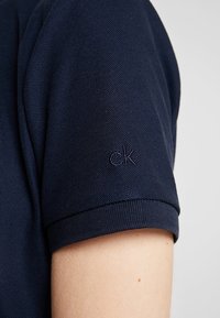 Navy blue polo shirt featuring a textured fabric with a fitted sleeve, detailed with an embroidered "ck" logo on the cuff.