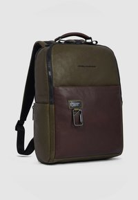 Rucksack mit einem oberen Teil aus grünem Leder und einem unteren Teil in Dunkelbraun, ausgestattet mit einem Reißverschluss, einem robusten Griff und einem Metalldetail vorne.