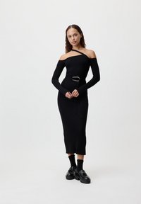 Crna rebrasta pletena haljina s off-shoulder dizajnom, dugim rukavima, remenom u struku i suptilnim prorezom na rubu, uparenim s robusnim crnim loafericama.