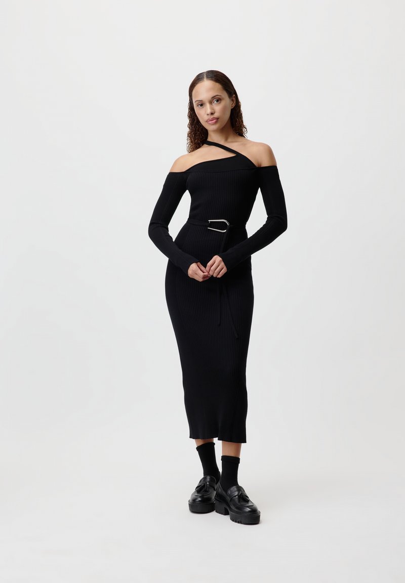 Crna rebrasta pletena haljina s off-shoulder dizajnom, dugim rukavima, remenom u struku i suptilnim prorezom na rubu, uparenim s robusnim crnim loafericama.