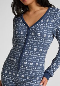 Body à manches longues bleu en tissu doux, orné de motifs de flocons de neige et de cœurs blancs, avec un décolleté en V profond et des détails de boutons.