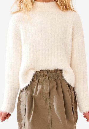 Frau trägt einen cremefarbenen, flauschigen Pullover, der in einen hoch taillierten, braunen Rock mit Knöpfen und Taschen gesteckt ist, dargestellt von Hals bis Mitte Oberschenkel.