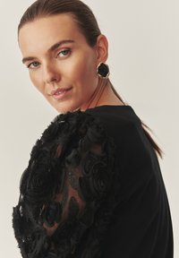 Fekete felső texturált fekete virágos csipkés ujjal; az modell arany és fekete nyolcszögletű füleket visel. Lágy, elegáns kialakítás és gazdag részletek.