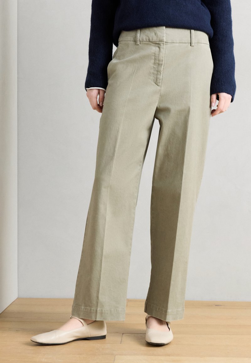 Άτομο που φοράει μπεζ παντελόνι φαρδιάς γραμμής, μπλε ναυτικό πουλόβερ και κρεμ επίπεδα παπούτσια, στέκεται σε φωτεινό ξύλινο πάτωμα μπροστά σε απλή λευκή τοίχο.