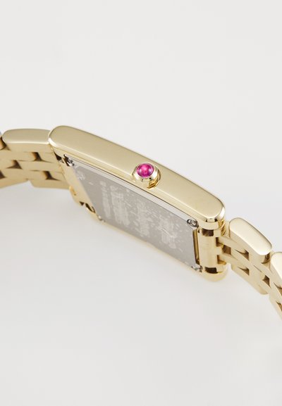 Montre dorée de forme rectangulaire, avec un bracelet en métal, un boîtier plat, et une pierre précieuse rose sur la couronne. Le dos est en argent avec des détails gravés.