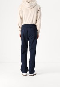 Beige hoodie med en bakficka, marinblå byxor med elastisk midja och vita sneakers med bruna sulor, mot en enkel vit bakgrund.