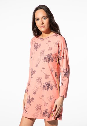 Frau mit langen dunklen Haaren trägt ein pinkfarbenes langärmeliges Kleid mit schwarzen botanischen und Insektenmotiven und steht vor einem weißen Hintergrund.