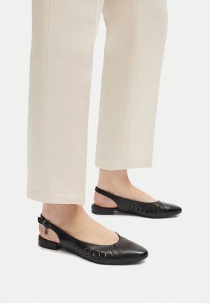 Zapatos slingback de cuero negro con detalles recortados y un tacón bajo y ancho. Combinados con pantalones de pierna recta de color crema.