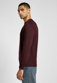 Burgundowy sweter z miękkiego materiału z okrągłym dekoltem i ściągaczami na mankietach. Przedstawiony w profilu bocznym na jasnym tle.