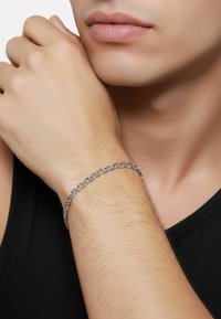 Bracciale a catena d'argento al polso, con maglie intrecciate e finitura liscia. Tono della pelle visibile, su sfondo di canottiera nera.