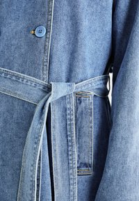 Giacca di jeans in azzurro chiaro, con vita legata, chiusura con bottoni blu e cuciture gialle a contrasto lungo i bordi e le tasche.
