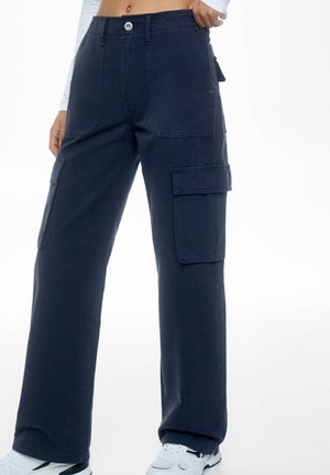 Marineblauwe wijde cargobroek met voor- en zijflappakken, gedragen met witte sneakers en een korte witte top met lange mouwen.