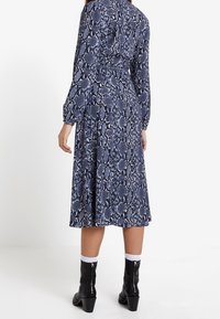 Kleid aus marineblauem Schlangenlederdruck mit langen Ärmeln, tailliertem Schnitt und ausgestellt Rock. Kombiniert mit schwarzen Stiefeletten und weißen Socken.