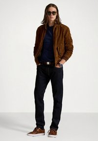 Hnědá semišová bunda, námořnicky modré tričko, tmavé džíny, hnědé volnočasové boty a zelené sluneční brýle; hodinky na zápěstí; volný styl; model stojí.