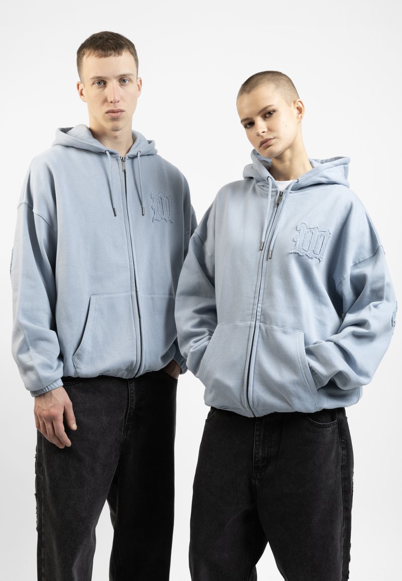 Sweatshirts bleu clair avec fermeture éclair à l'avant, grandes poches et détails de logo brodés, associés à un jean noir à jambes larges. Design simple et coupe décontractée.