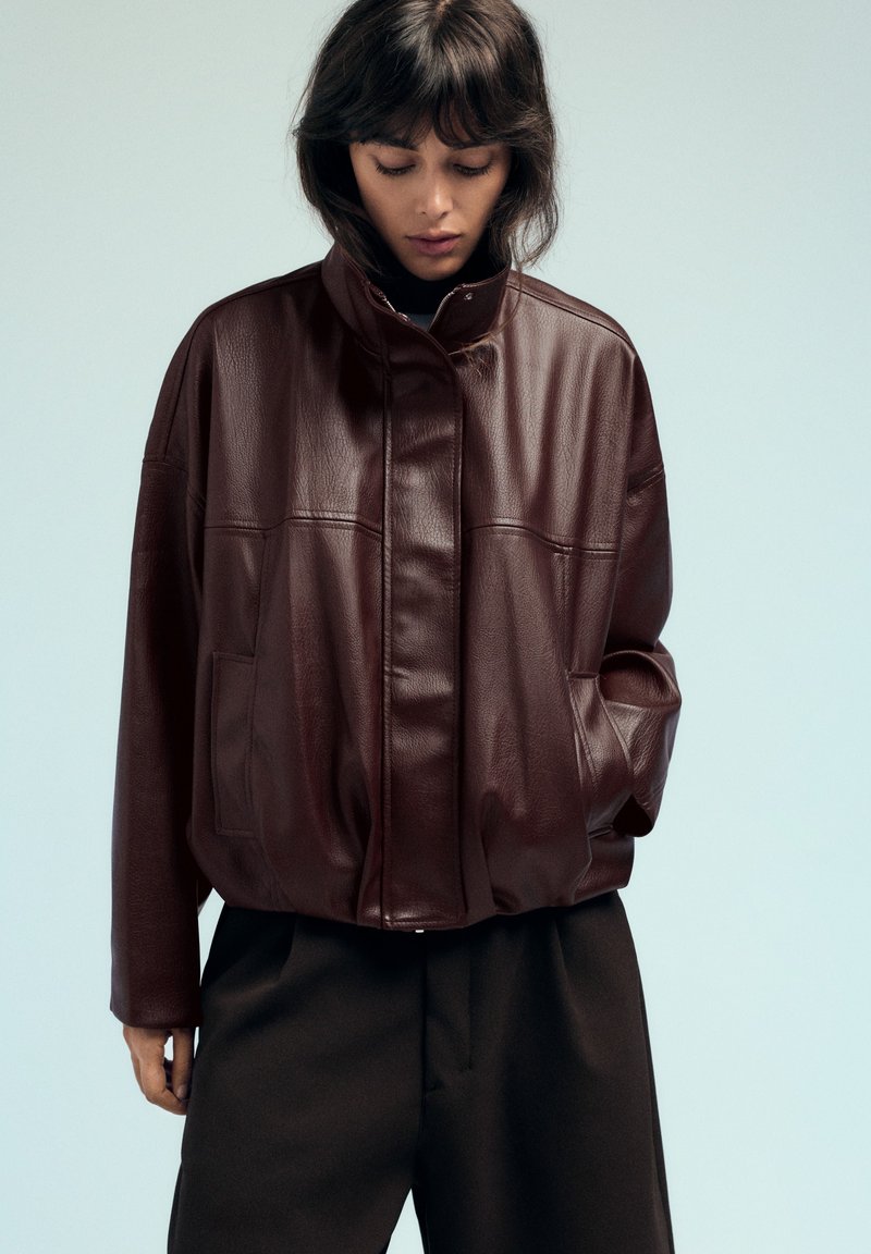 Femme portant une veste en cuir marron foncé avec un col montant et un pantalon noir, regardant vers le bas, une main dans la poche de la veste.