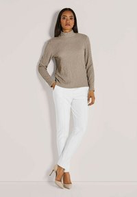 Frau steht an einer hellen Wand und trägt ein strukturiertes beiges Langarmoberteil, weiße enganliegende Hosen und beige High Heels.