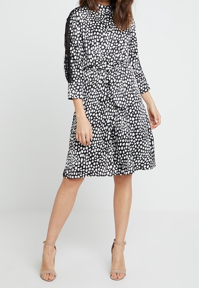 Femme portant une robe mi-longue noire à pois blancs avec des manches trois-quarts et des sandales à talons hauts beiges avec bride à la cheville.