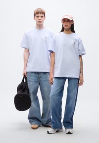 Δύο μοντέλα φορούν ανοιχτό μωβ μπλουζάκια με μικρά λογότυπα. Τα συνδυάζουν με τζιν παντελόνια φαρδιάς γραμμής. Το ένα κρατά μια μαύρη στρογγυλή τσάντα, το άλλο φορά ένα ροζ καπέλο.