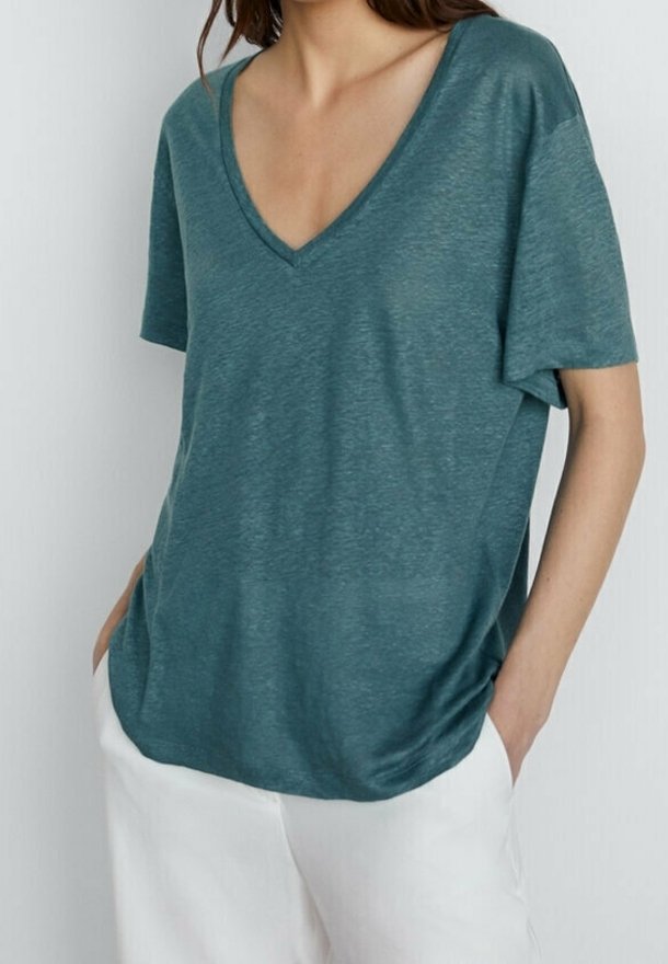 Femme portant un t-shirt ample bleu sarcelle à col en V et manches courtes avec un pantalon blanc, mains dans les poches, sur un fond clair uni.
