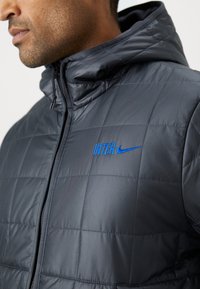 Izolirana tamno siva puffer jakna s kapuljačom, koja ima plavi "INTER" logo i Nike oznaku na grudima. Glatka, sjajna tekstura materijala.