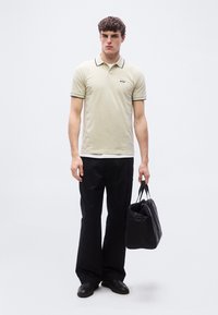 Jeune homme portant un polo beige, un pantalon noir, des chaussures noires, tenant un sac noir, debout devant un fond blanc uni.