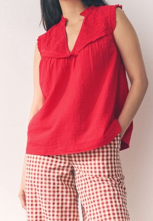 Femme portant un chemisier rouge sans manches avec un col brodé et un pantalon à carreaux rouges et blancs, une main dans la poche, sur fond uni.