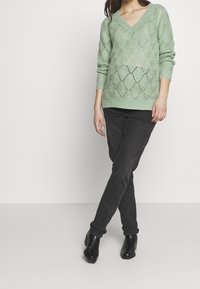 Pull en maille vert menthe avec un col en V, présentant un motif en losange. Associé à un jean noir ajusté et des bottines noires.