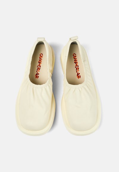 White slip-on shoes with a soft fabric upper, elasticated opening, and a contrasting red logo inside the shoe. Round toe and flat sole.