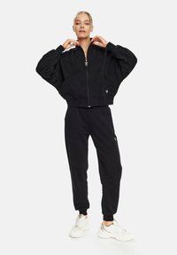 Crna skraćena bomber jakna s oversized rukavima, zatvaranjem na zip, i bočnim džepovima, u kombinaciji s odgovarajućim jogger hlačama koje sadrže detalj s logotipom.