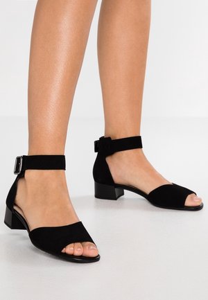 Sandales ouvertes en suede noir avec un petit talon bloc, dotées d'une bride à la cheville maintenue par une boucle en argent. Texture lisse et mate.