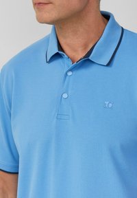 Light blue polo shirt made of textured fabric, featuring a dark blue collar, three buttons, and a logo on the left side.
