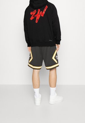 Osoba stojąca tyłem ubrana w czarną bluzę z kapturem z czerwonym nadrukiem, szare spodenki do koszykówki z żółtym obszyciem, białe skarpety sportowe i białe tenisówki.