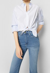 Camisa de botones blanca y azul claro a rayas atada en la cintura, combinada con jeans azul claro. La camisa tiene cuello y mangas enrolladas.