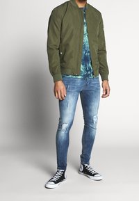 Man in een groene bomberjack, een blauwe tie-dye shirt, versleten skinny jeans en zwarte high-top sneakers, staande tegen een effen achtergrond.