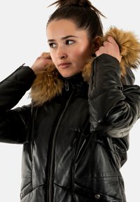 Veste matelassée en cuir noir avec capuche amovible doublée de fourrure, fermeture éclair et poches latérales. Texture lisse, design matelassé avec coutures.