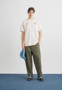 Oversized ανοιχτό ροζ μπλούζα με γραφικά, συνδυασμένη με ελαιδί cargo παντελόνια. Ένα μπλε καπέλο κρατιέται στο αριστερό χέρι· φορούνται μαύρα αθλητικά παπούτσια.