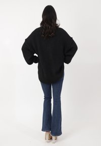Suéter negro de gran tamaño con un acabado texturizado, ajuste holgado y hombros caídos, combinado con jeans acampanados azules y zapatos de tacón alto.