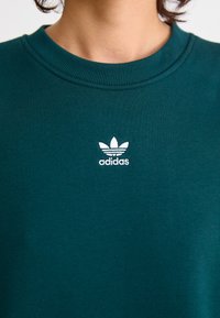 Gros plan sur un sweat-shirt à col rond vert avec un petit logo trèfle blanc Adidas centré sur la poitrine.