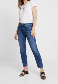 Modré džíny z denimu se střihovým střihem, s pěti kapsami a mírným vyblednutím. V páru s bílým tričkem a černými sandály na podpatku.